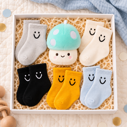 A gift set of baby socks and a plush toy arranged in a white box with crinkle paper filler. The set includes gray, cream, black, yellow, and light blue socks, each with a black smiley face, plus a mint green and white mushroom plush toy.