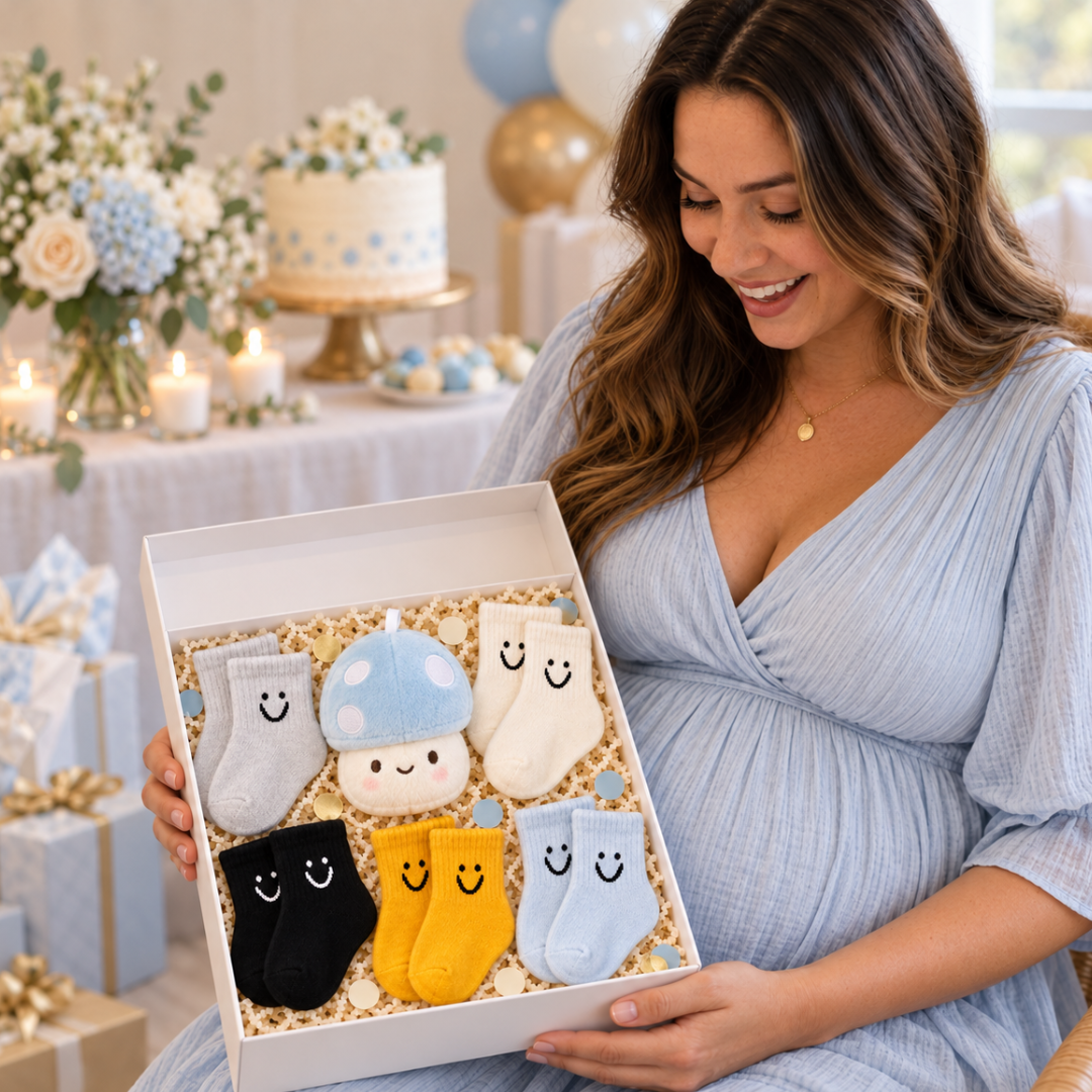 A pregnant woman in a light blue dress holds an open gift box filled with six cute baby socks in different colors, including gray, light blue, cream, black, yellow, and light blue. The background shows a decorated baby shower or birthday party with a cake, candles, and balloons.
