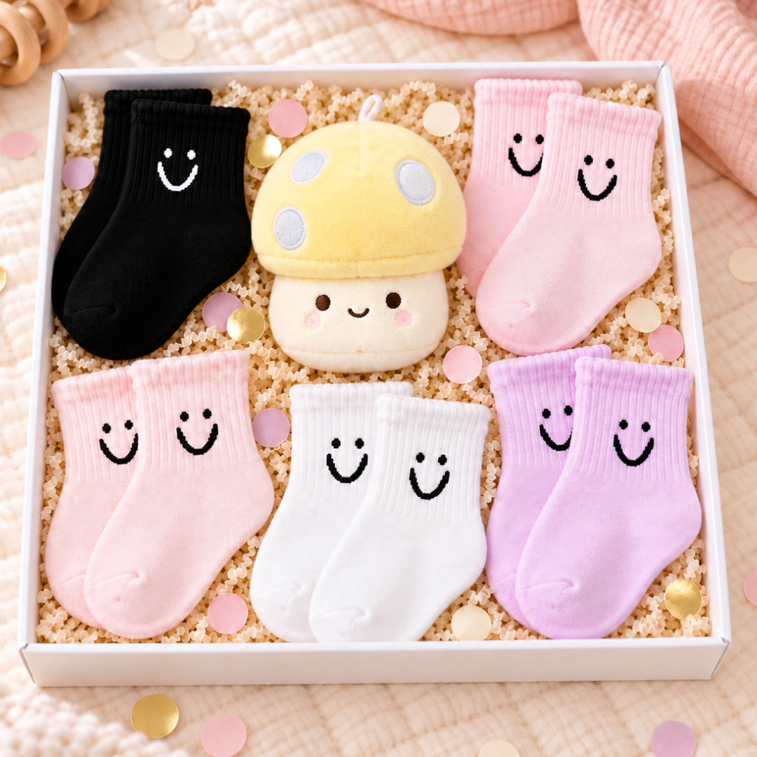 A gift box of baby socks and a plush toy arranged in a white box filled with beige paper shred and confetti. The set includes a black pair, a yellow mushroom plush, and four pastel pink, white, and lavender pairs of socks, all with embroidered smiley faces.