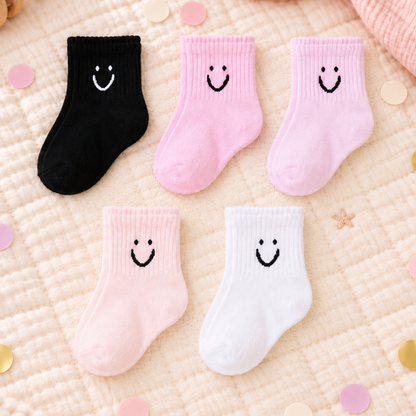 Five pairs of baby socks arranged on a quilted cream blanket with scattered confetti. The socks come in black, light pink, medium pink, white, and pale pink, each with a simple embroidered smiley face on the front.