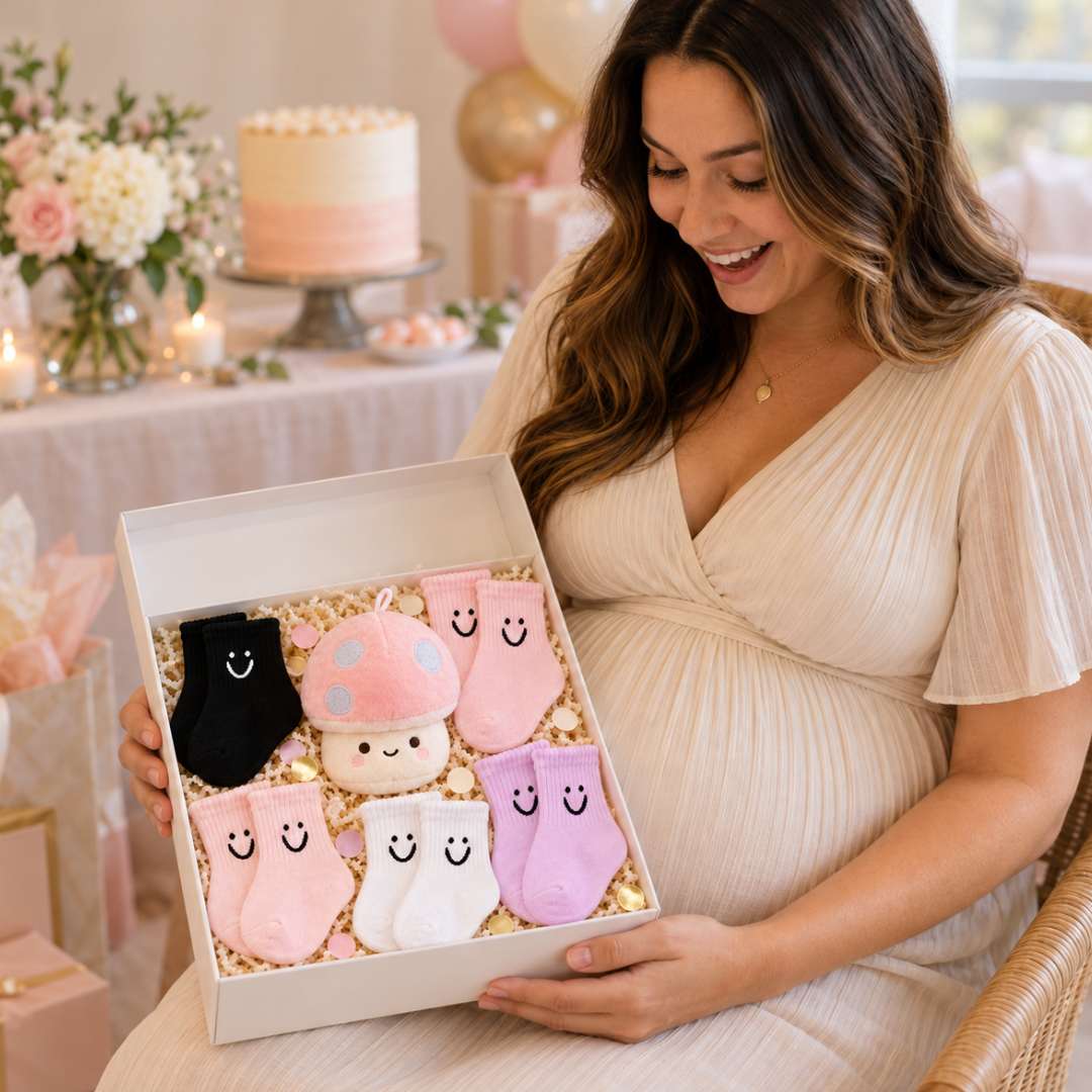 A pregnant woman in a cream dress holds an open gift box filled with baby socks. The socks are in pastel pink, lavender, white, and black, decorated with cute cartoon faces.