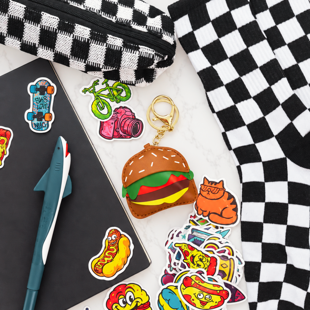 A collection of colorful die-cut stickers displayed on a white marble surface alongside a black and white checkered pencil case, a notebook, a shark-shaped pen, and a plush toy keychain shaped like a hamburger with a gold key ring.