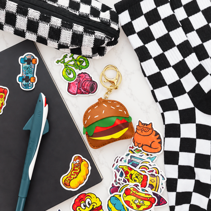A collection of colorful die-cut stickers displayed on a white marble surface alongside a black and white checkered pencil case, a notebook, a shark-shaped pen, and a plush toy keychain shaped like a hamburger with a gold key ring.