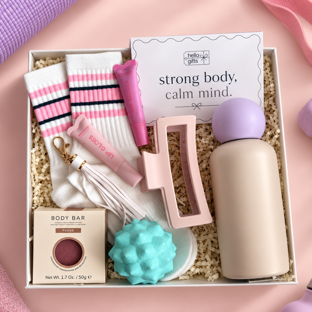 A curated gift box set from Hello Girls featuring a body bar, lip gloss, socks, a pink stand, a purple ball, a teal textured sponge, and a beige bottle, arranged in shredded paper on a pink background.