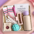 A curated gift box set from Hello Girls featuring a body bar, lip gloss, socks, a pink stand, a purple ball, a teal textured sponge, and a beige bottle, arranged in shredded paper on a pink background.