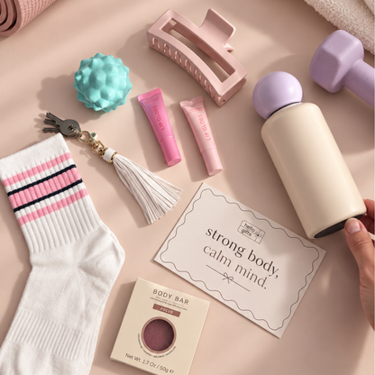 A styled flat-lay of wellness and self-care items on a soft blush background, including a cream bottle with a lavender top held by a hand, pink lip balm tubes, a pink hair claw clip, a white sock with pink and black stripes, a small teal massage ball, keys with a tassel, a body bar package, and a card reading &