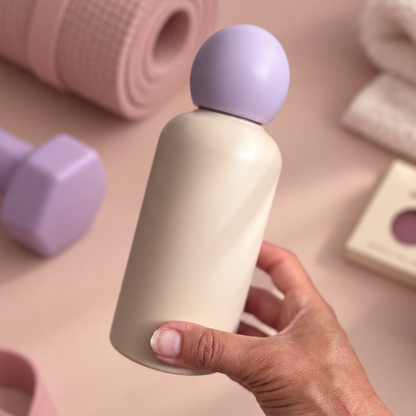 A hand holding a matte cream-colored bottle with a lavender spherical cap, set against a soft pink background with blurred fitness props.