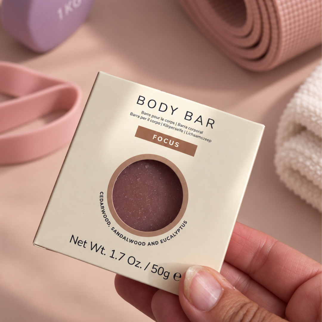A hand holding a square cream-colored box of Focus Body Bar, showing the circular window that reveals the textured brown body bar inside. The background is a soft beige with blurred pink and white wellness props.