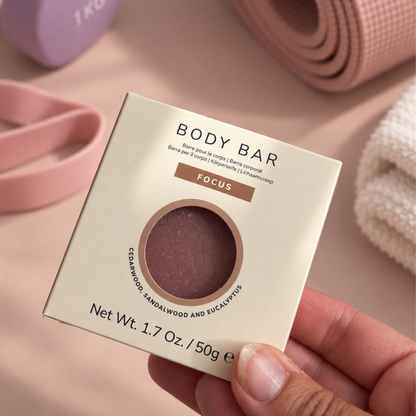 A hand holding a square cream-colored box of Focus Body Bar, showing the circular window that reveals the textured brown body bar inside. The background is a soft beige with blurred pink and white wellness props.