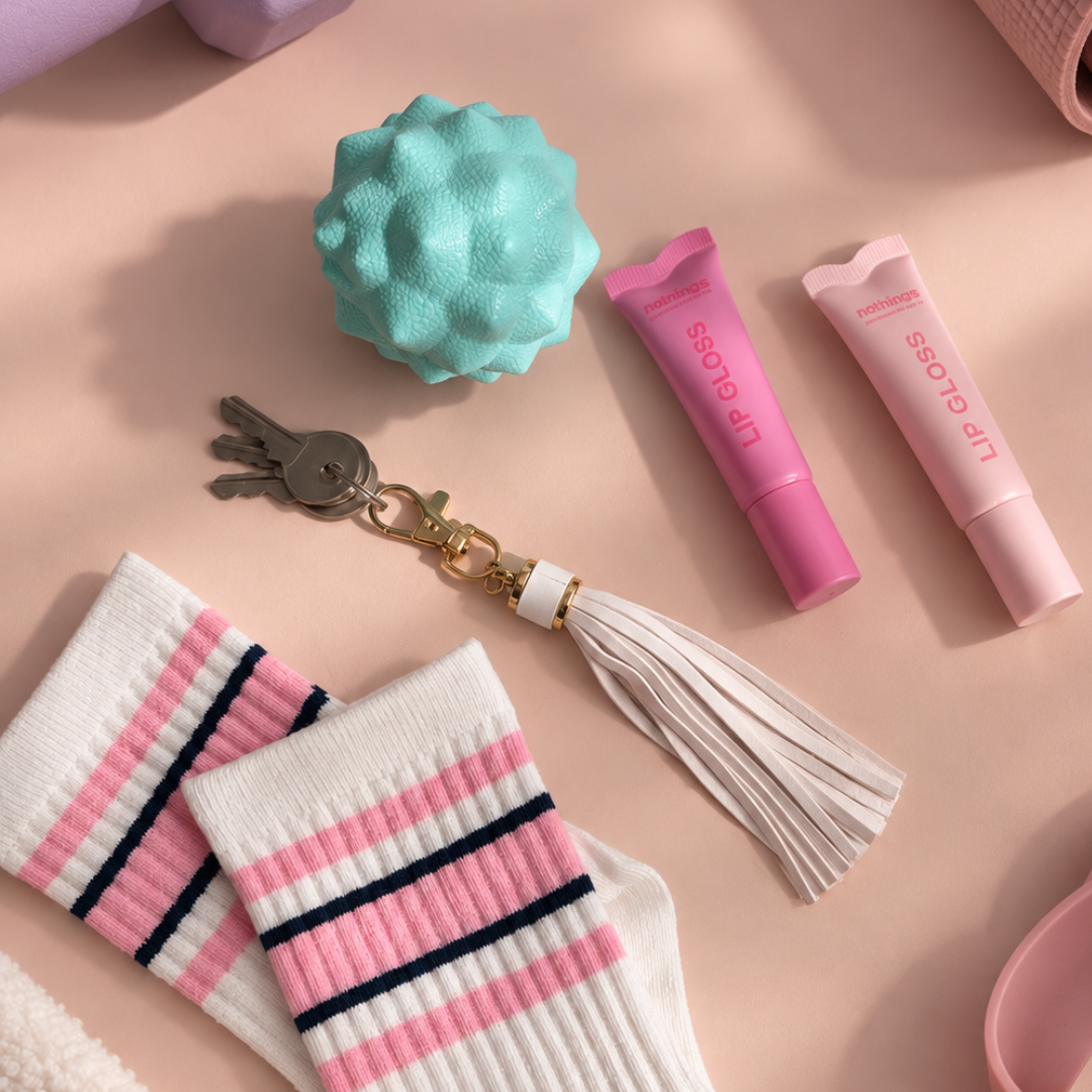 A styled flat-lay featuring a pair of white knit socks with pink and navy stripes, a mint green textured ball, a pink lip gloss tube, a pale pink lip gloss tube, and a white tassel keychain with gold hardware, all arranged on a soft pink background.