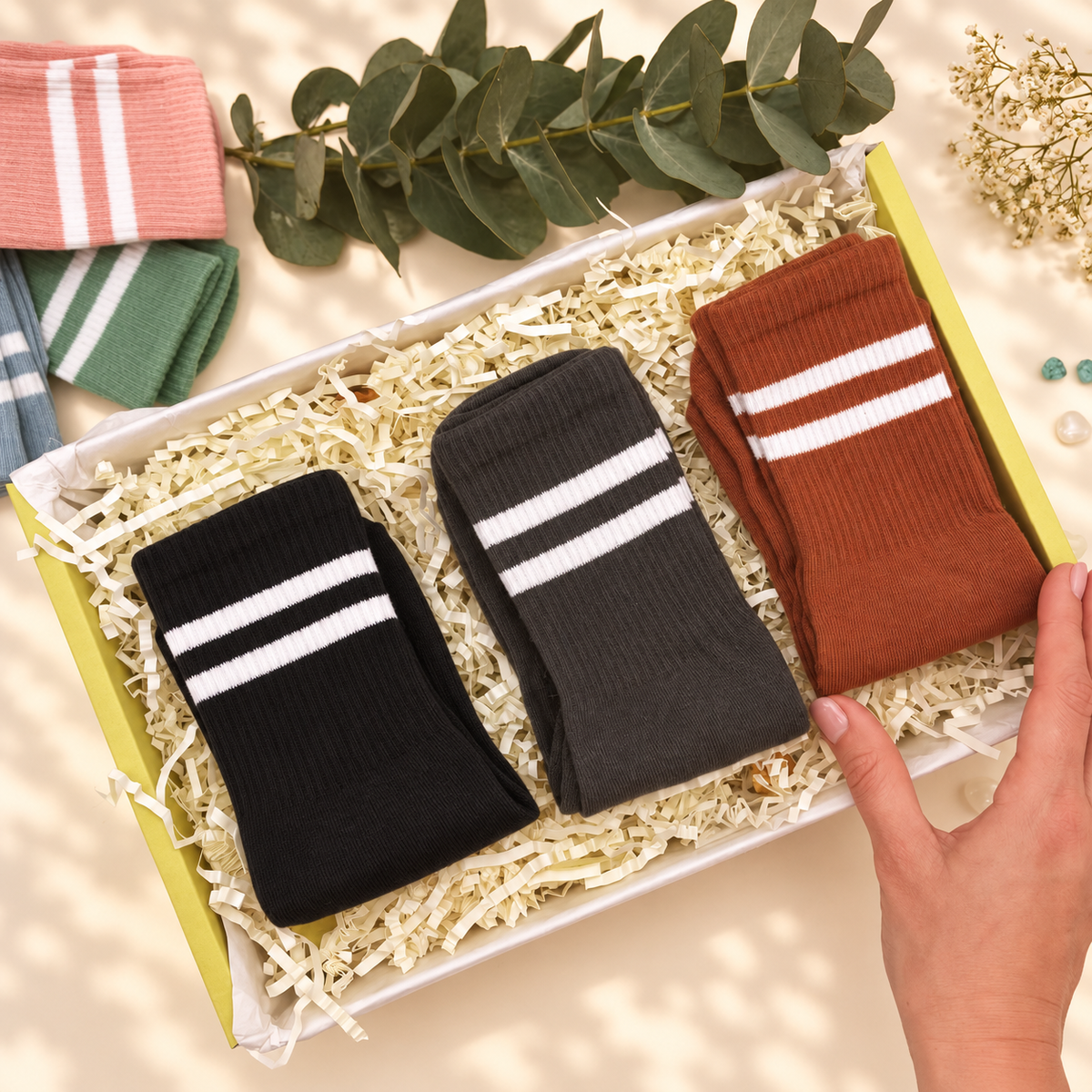 Three pairs of ribbed crew socks with white stripes are displayed in a gift box filled with shredded paper. A hand is holding the box, and decorative greenery and folded socks are arranged around it on a warm beige surface.