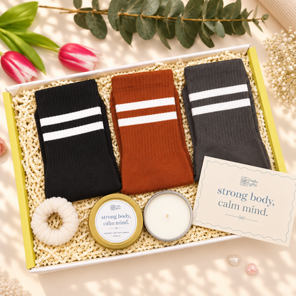 A gift box set containing three pairs of socks in black, rust orange, and charcoal gray with white stripes, a white scrunchie, a gold tin candle, a white candle, and a card that reads &