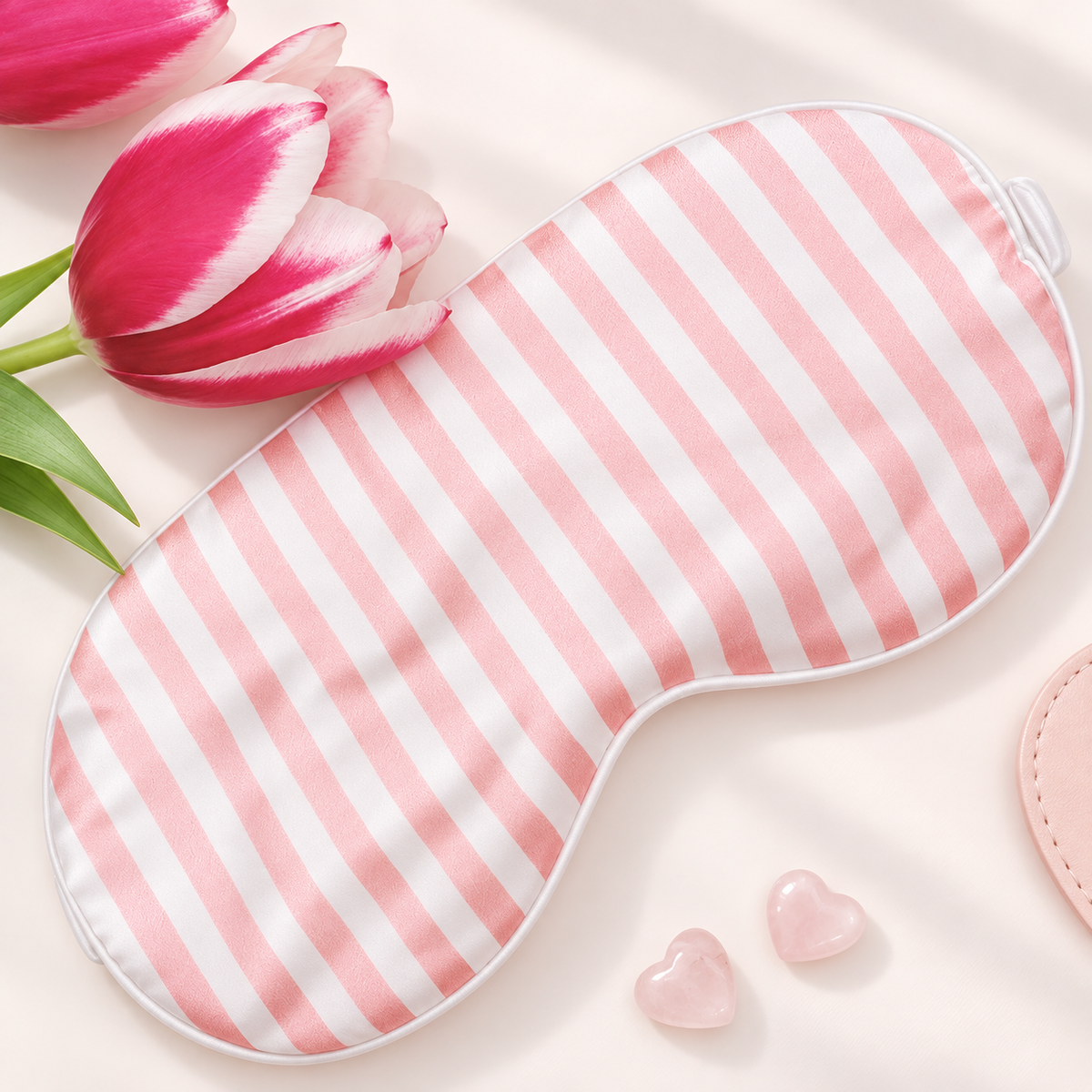 Pink and white striped sleep mask with a white border, styled on a light cream background with pink tulips and two small pink heart-shaped stones.