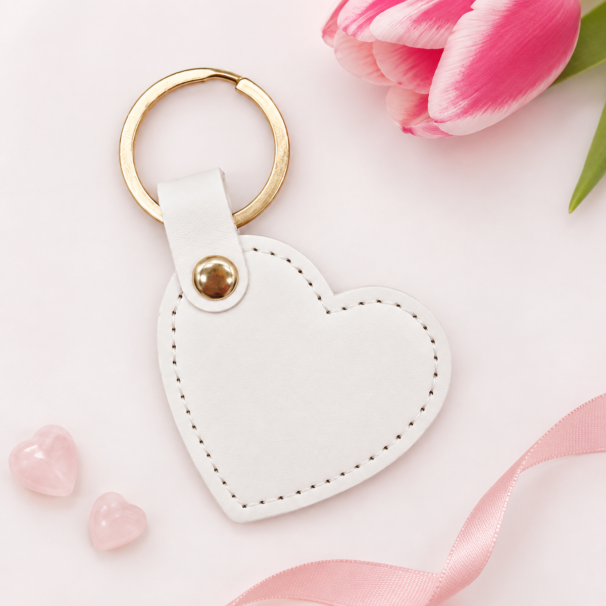 A white faux-leather heart-shaped keychain with a gold-tone split ring and snap button, styled on a white background with a pink tulip, pink stones, and a pink ribbon.