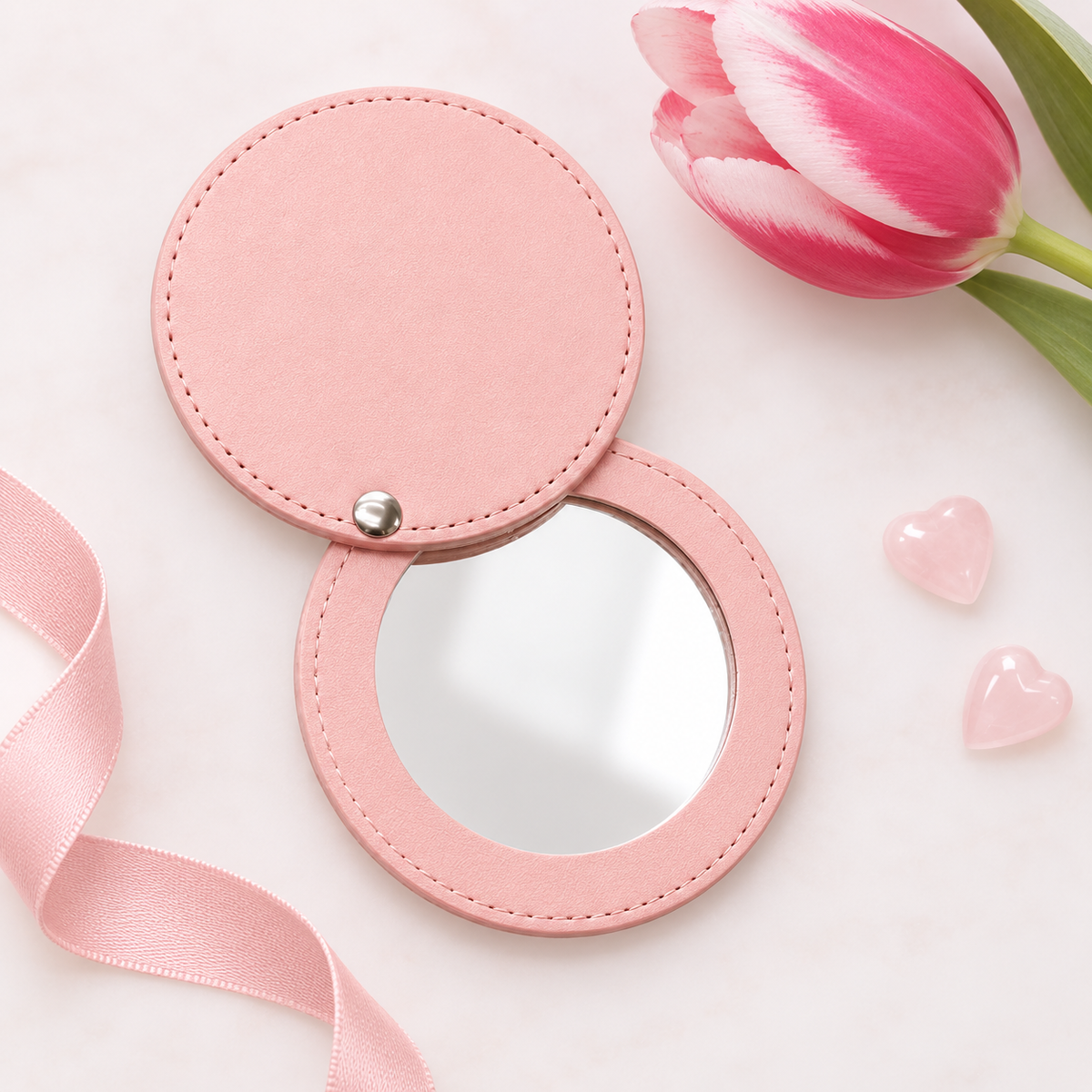 A pink round folding compact mirror with a textured faux-leather cover, shown open on a white marble background with a pink tulip, pink ribbon, and two small pink heart charms.