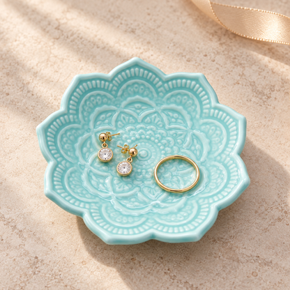 A light aqua ceramic jewelry organizer tray with a scalloped edge and embossed floral pattern, holding a pair of gold hoop earrings and a gold ring, set on a textured beige surface with soft shadows.