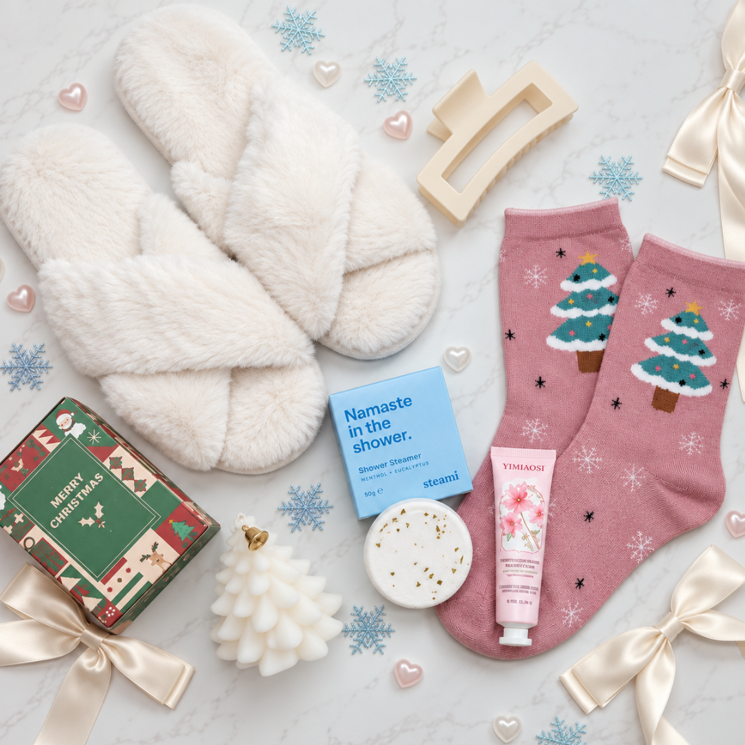 A styled flat-lay of spa and holiday gift items on a light marble surface, including fluffy white slippers, pink socks with Christmas tree motifs, a wrapped gift box, a blue soap bar labeled &