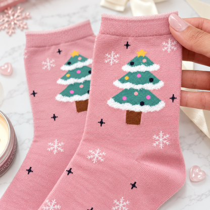 Pink crew socks with a Christmas tree and snowflake pattern, held by a hand over a marble surface with holiday decorations.