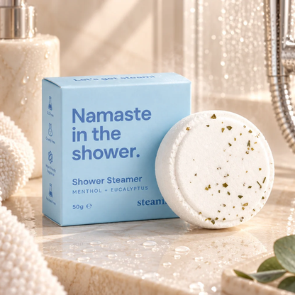 A light blue box of Namaste in the Shower steamers next to a round white bath steamer disc with dried herb specks, set on a marble bathroom counter with a shower head and soap dispenser in the background.