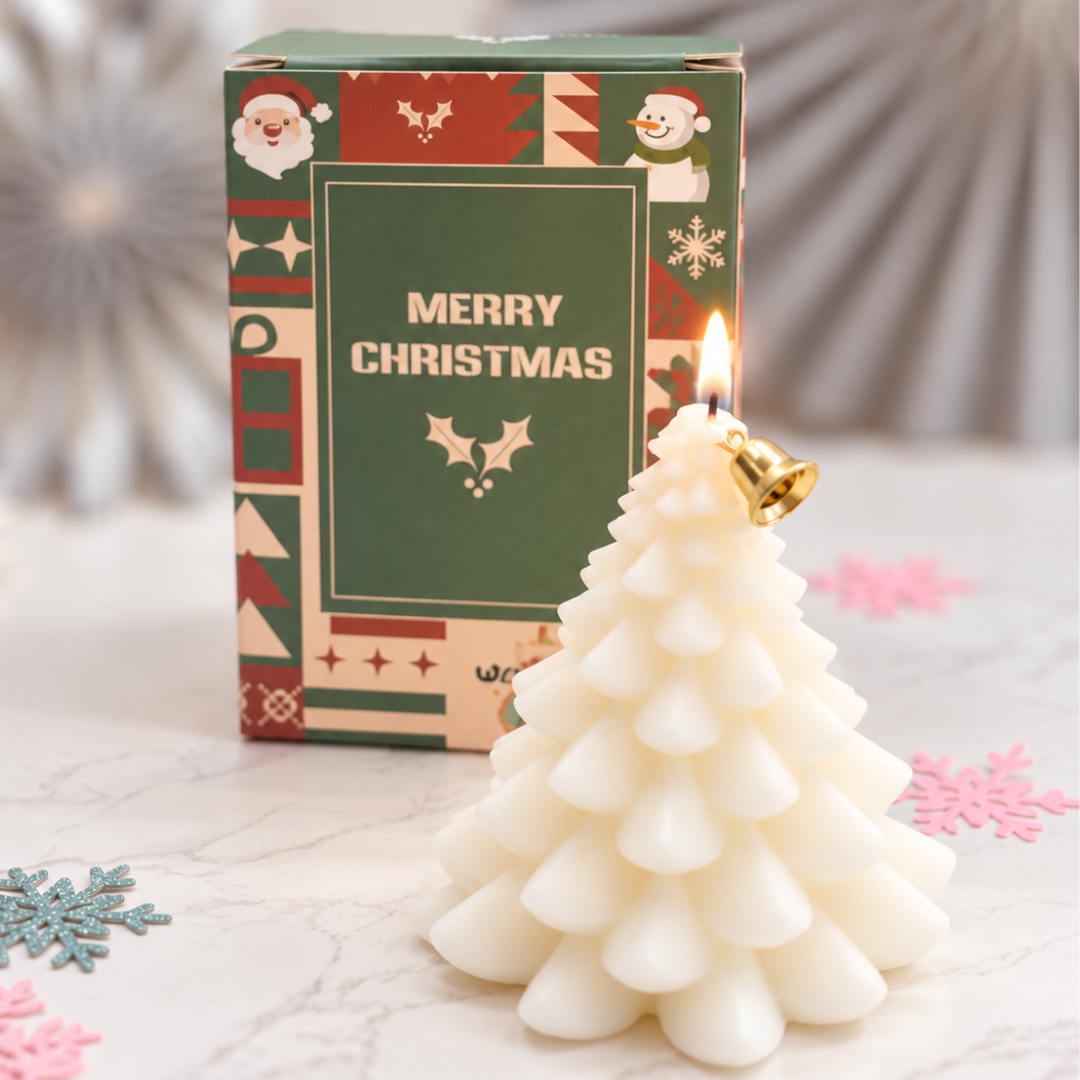 A Christmas tree-shaped white candle with a small gold bell detail and a visible flame, placed in front of its green and red patterned Christmas box on a light marble surface.