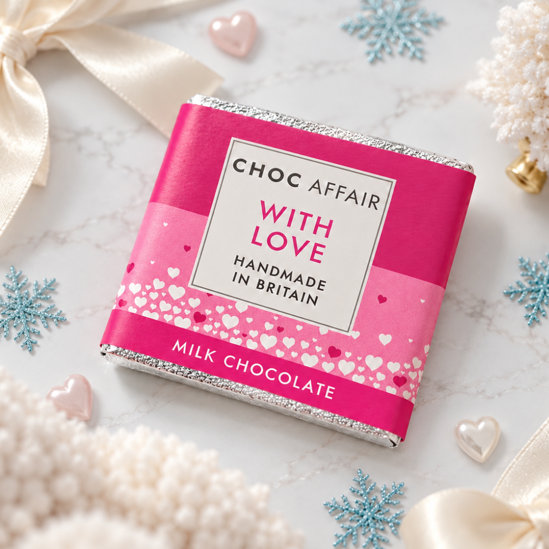 A milk chocolate bar wrapped in a bright pink paper sleeve with silver foil edges, photographed on a light marble surface with decorative white ribbons, snowflake ornaments, and small heart props. The label reads &