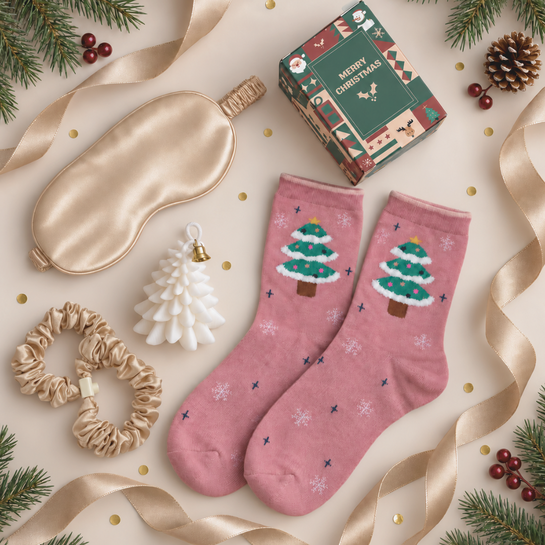 A styled flat-lay of a Christmas-themed gift set on a beige background with gold ribbons, pine branches, and gold confetti. The set includes a pair of dusty pink crew socks with green and white Christmas tree designs, a champagne gold satin sleep mask, a white mini Christmas tree ornament, a beige scrunchie, and a green &