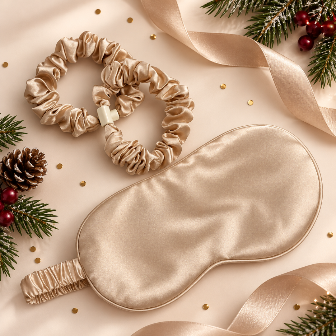A beige satin sleep mask and matching scrunchie are arranged on a warm cream background with ribbon, pine branches, berries, and small gold confetti accents.