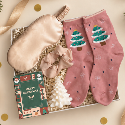 A Christmas-themed gift set in a white box filled with shredded paper, featuring dusty pink socks with embroidered green Christmas trees, a champagne satin sleep mask, a matching scrunchie, a small white Christmas tree candle, and a green &