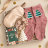 A Christmas-themed gift set in a white box filled with shredded paper, featuring dusty pink socks with embroidered green Christmas trees, a champagne satin sleep mask, a matching scrunchie, a small white Christmas tree candle, and a green &