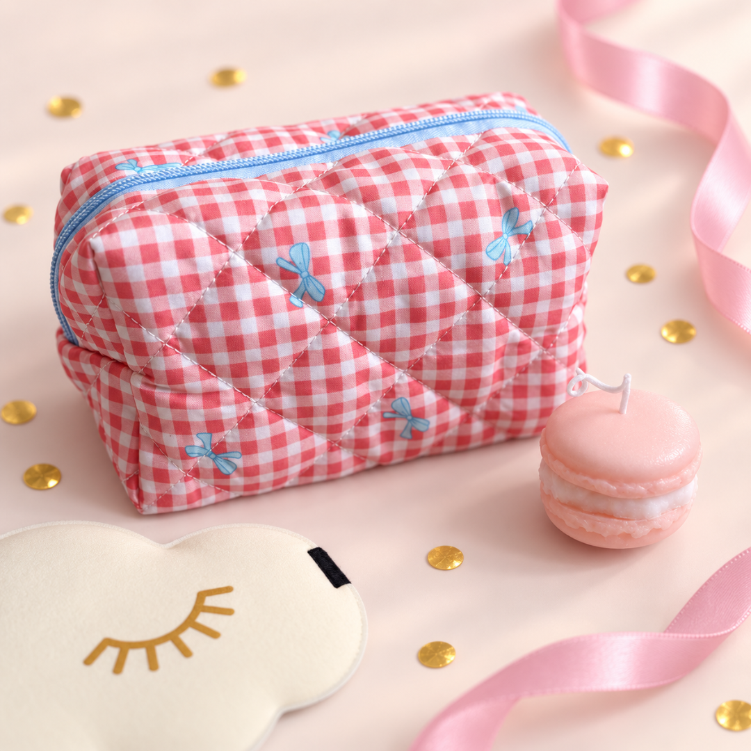 A pink and white gingham quilted cosmetic pouch with blue zipper and embroidered bows, styled on a light pink background with a macaron, eye mask, and ribbon.