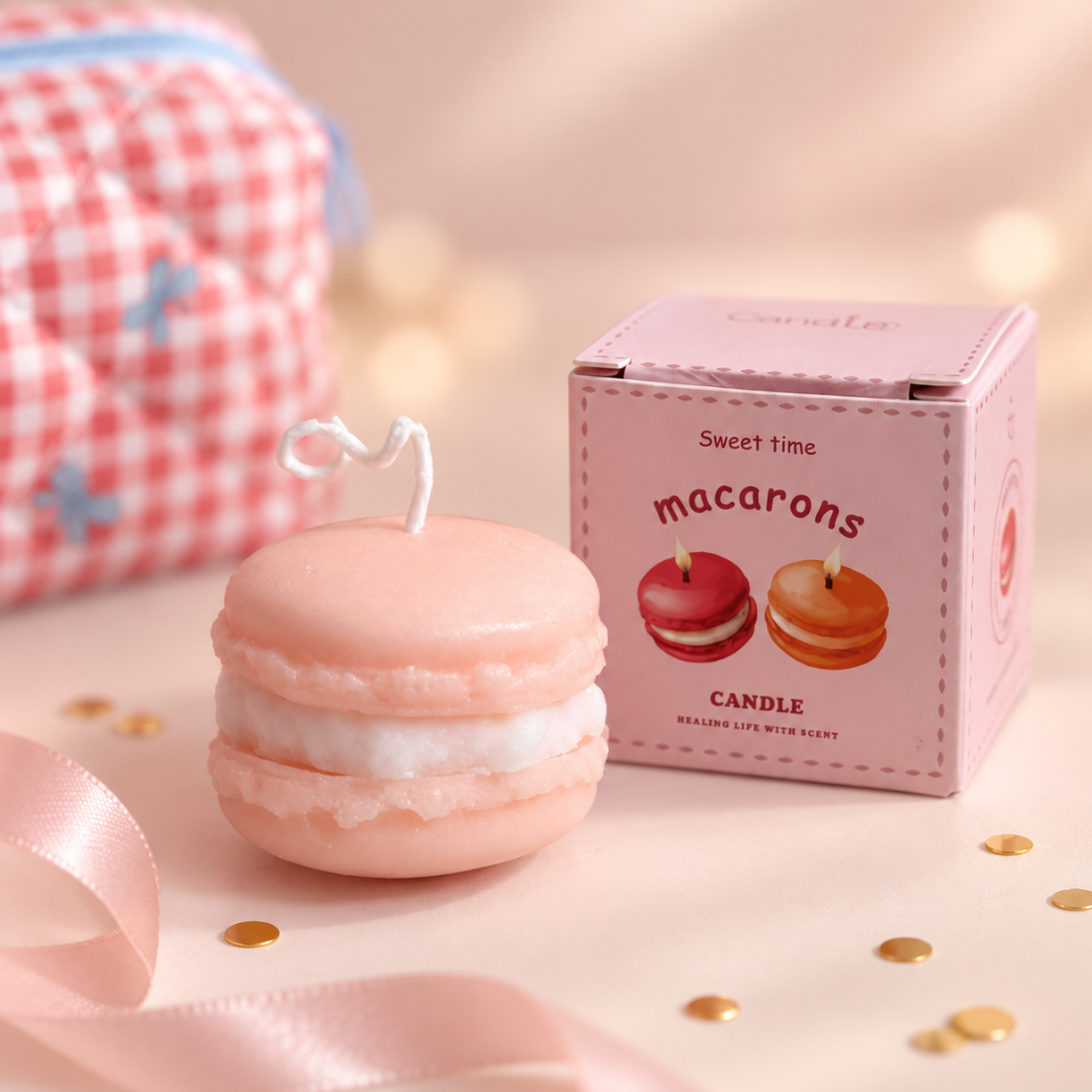 A pink macaron-shaped candle with a white cream filling and a small heart-shaped wick, shown next to its pink packaging box labeled &