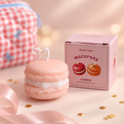 A pink macaron-shaped candle with a white cream filling and a small heart-shaped wick, shown next to its pink packaging box labeled &