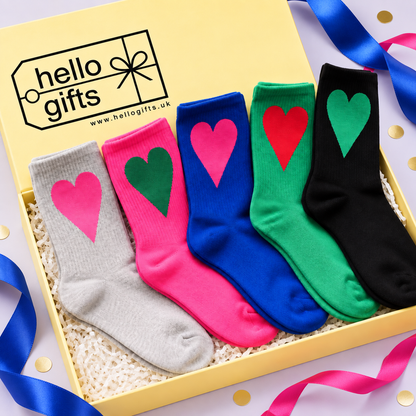 Five pairs of crew socks in gray, pink, blue, green, and black, each with a contrasting heart on the front, displayed inside a yellow Hello Gifts box with shredded paper filler and ribbon accents.