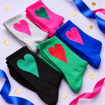 Six pairs of crew socks in black, gray, green, pink, blue, and another green pair, each decorated with a large solid heart in a different color, arranged on a light lavender background with blue and pink ribbons and gold confetti.
