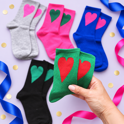 A hand holds a pair of green socks with red hearts, surrounded by other color variants (gray, pink, blue, black) and decorative ribbons and confetti on a lavender background.