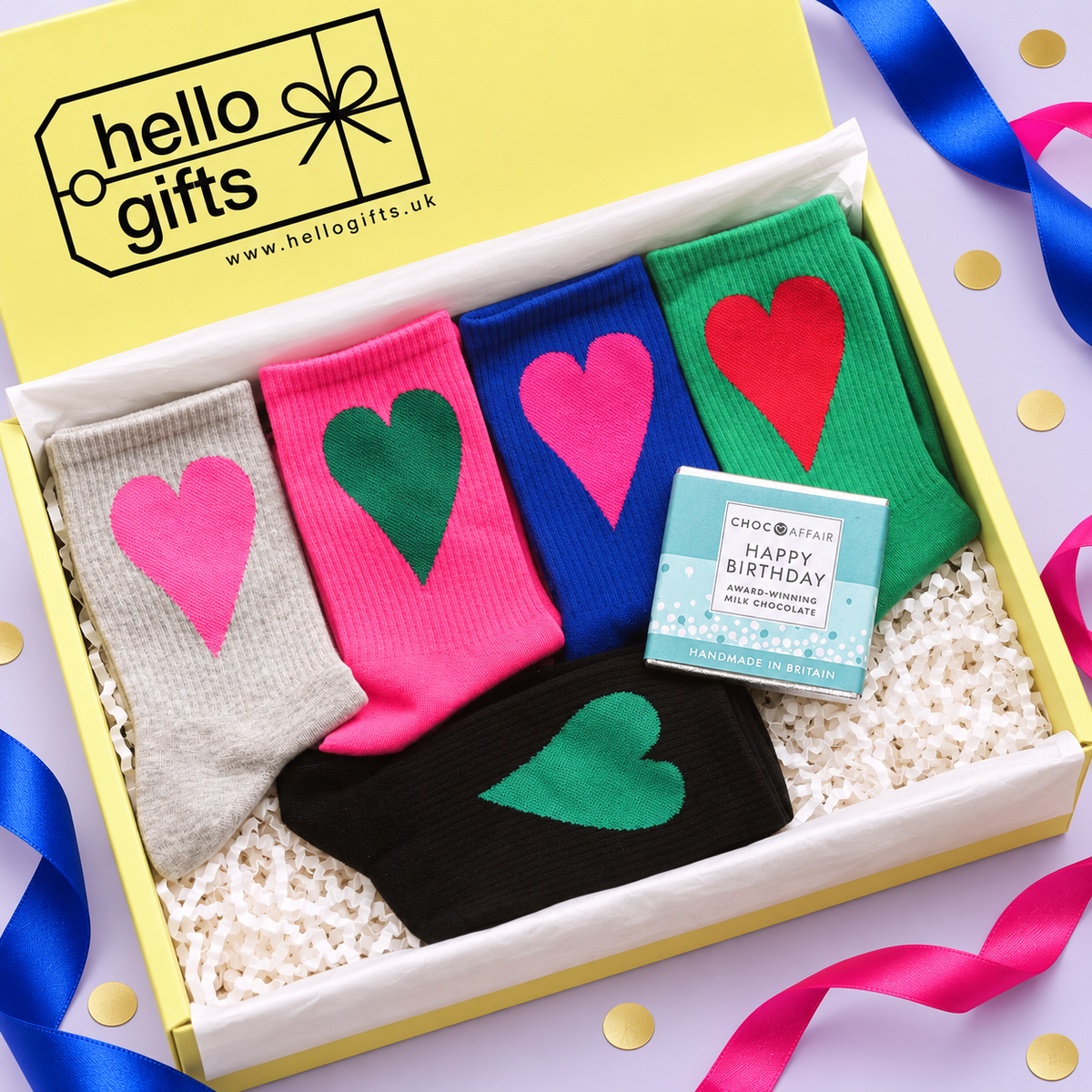 A gift box containing four pairs of socks in gray, pink, blue, and black, each with a large heart in a different color. A small chocolate bar and a Hello Gifts branded card are placed inside the box, which is set on a light purple surface with colorful ribbons and gold confetti.