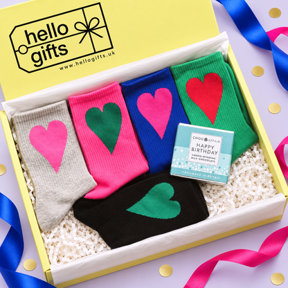 A gift box containing four pairs of socks in gray, pink, blue, and black, each with a large heart in a different color. A small chocolate bar and a Hello Gifts branded card are placed inside the box, which is set on a light purple surface with colorful ribbons and gold confetti.