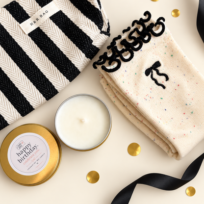 A styled flat-lay gift set featuring a black and white striped bag from HER BAG, a cream candle in a gold tin with a &