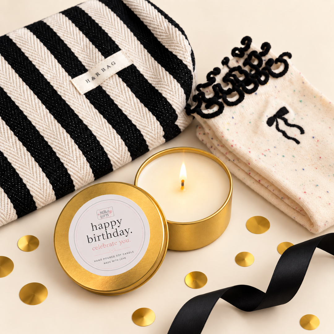 A curated gift set on a warm cream background featuring a black and white striped HEBRAGO pouch, a gold tin candle with a lit flame and white wax, a folded cream knit sock with black lace trim, gold confetti, a black ribbon, and a gold round sticker reading &