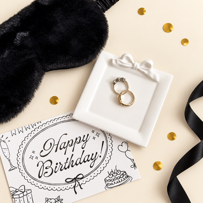 A styled flat-lay featuring a black plush blindfold, a white square tray holding two gold rings, a black ribbon, a black-and-white &
