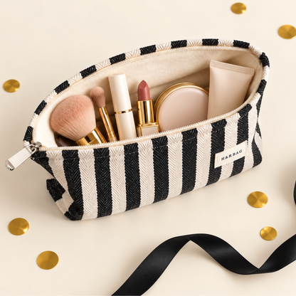 A striped cosmetic pouch with black and cream horizontal stripes, shown open with makeup items inside on a light beige background with scattered gold confetti and a black ribbon.