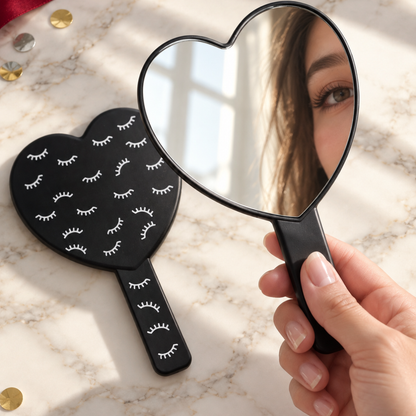A heart-shaped handheld mirror with a black handle and a black heart-shaped back decorated with white eyelash illustrations, shown on a marble surface with a hand holding the mirror and a reflection of a woman&