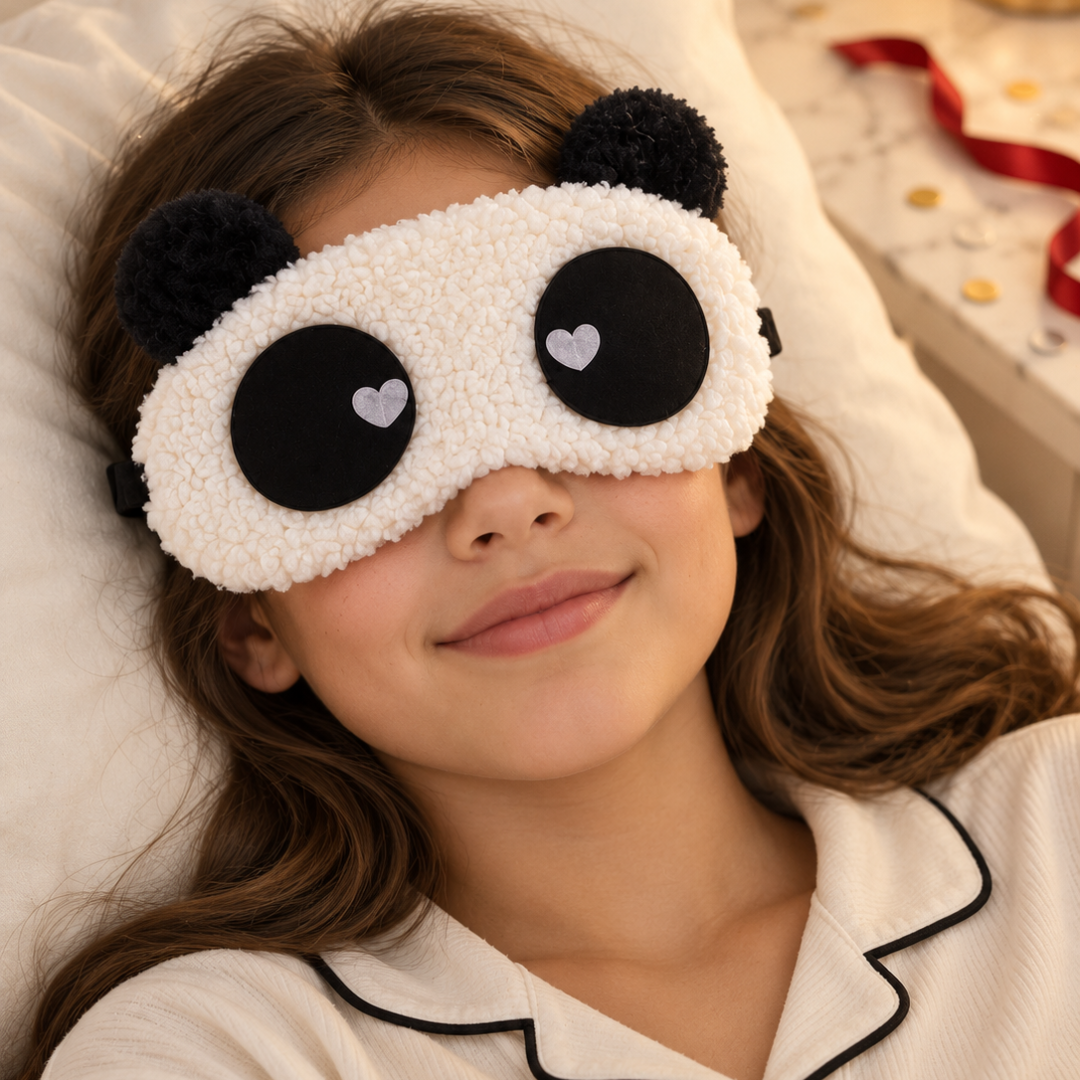 A child wearing a plush white and black panda-themed sleep mask, lying on a white pillow in a bedroom setting.