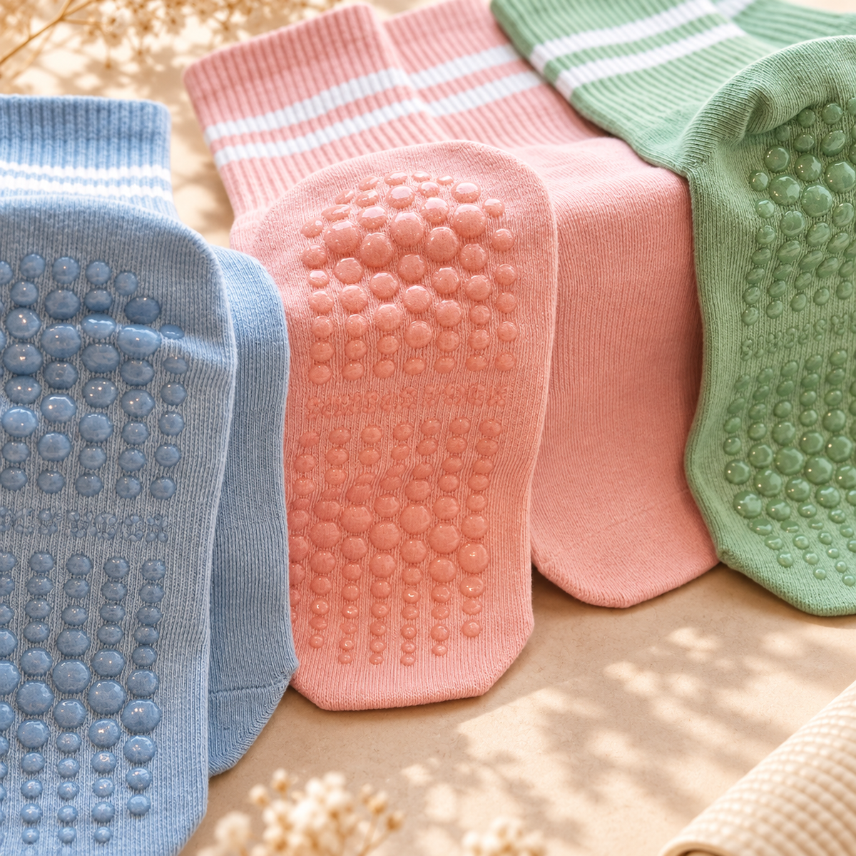 Four pairs of grip socks in pastel blue, pink, peach, and mint green, arranged overlapping on a beige surface with soft natural shadows.