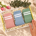 Three pairs of striped socks in pink, blue, and green are arranged in a gift box filled with shredded paper, with a hand holding the box edge. Decorative flowers, leaves, and a soft fabric surface surround the box.