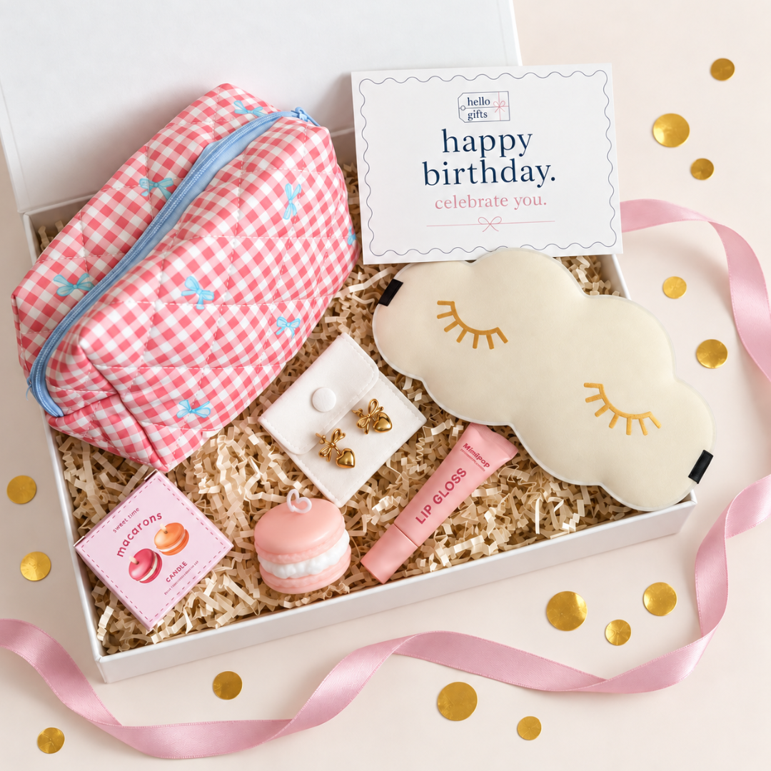 A curated gift box set containing a pink and white gingham makeup pouch, a cream cloud-shaped sleep mask with gold embroidered eyes, a pink macaron toy, a pink lip gloss tube, a small jewelry box, and a &