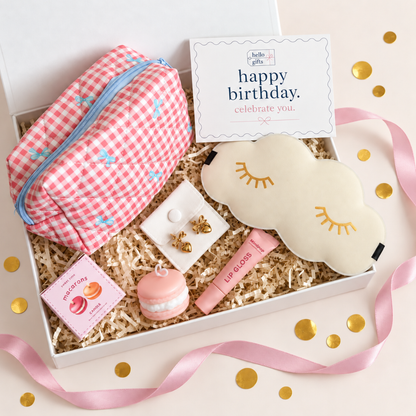 A curated gift box set containing a pink and white gingham makeup pouch, a cream cloud-shaped sleep mask with gold embroidered eyes, a pink macaron toy, a pink lip gloss tube, a small jewelry box, and a &