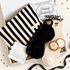 A curated gift box set featuring a black and cream striped bath towel, a black faux-fur sleep mask, cream speckled socks with black embroidery, a white candle in a gold tin, a small white gift box, and a &
