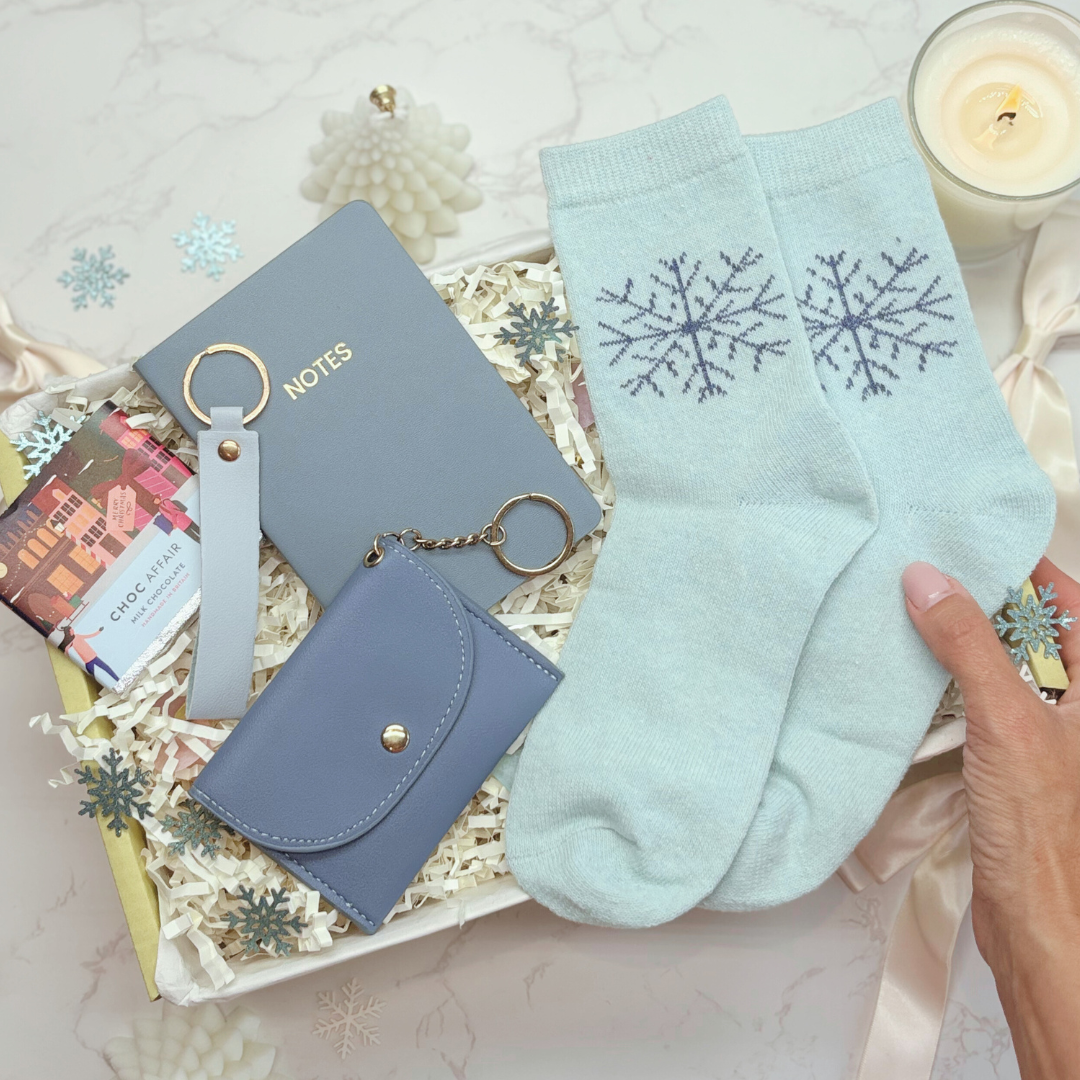 Light blue socks with snowflake design held by a hand, next to a notebook, wallet, and gift box on a decorative surface.