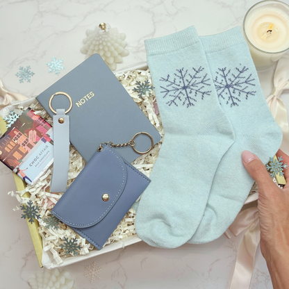 Light blue socks with snowflake design held by a hand, next to a notebook, wallet, and gift box on a decorative surface.
